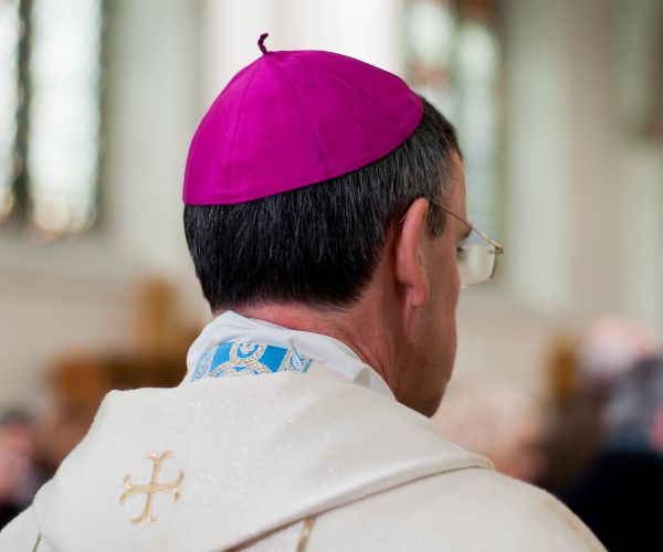 Camera captures the back and profile of Bishop John Arnold as he enters Salford Cathedral. He is wearing white vestments and his amaranth zucchetto
