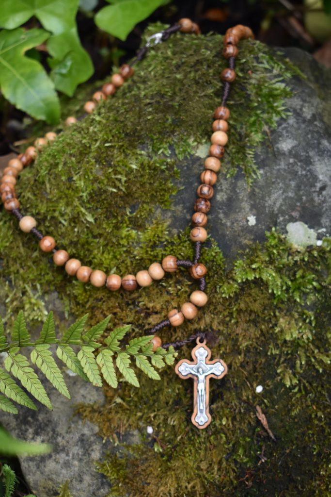 A wooden Rosary rests on a mossy rock, surrounded by ferns