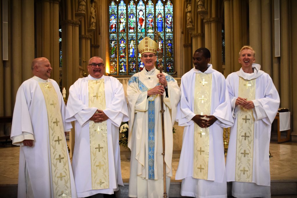 Deacon Lee on the far left is joined by his brother deacons and Bishop John in front of the stained glass window of the cathedral 