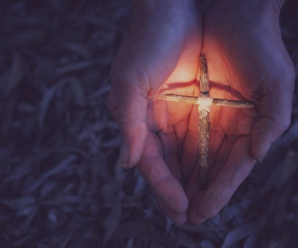 Pictured shows a small, wooden cross held in someone hands, surrounded by light in the darkness