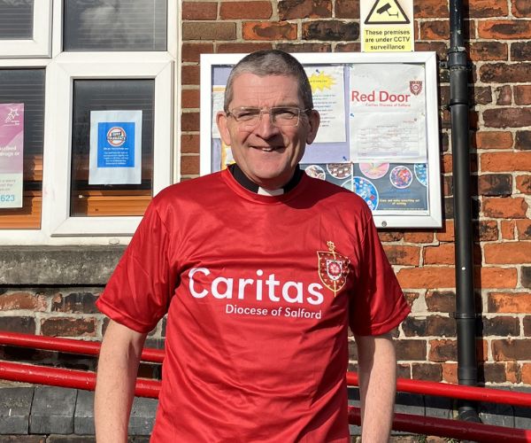 Fr Chris Gorton stands in front of Red Door wearing a red Caritas Salford sports top