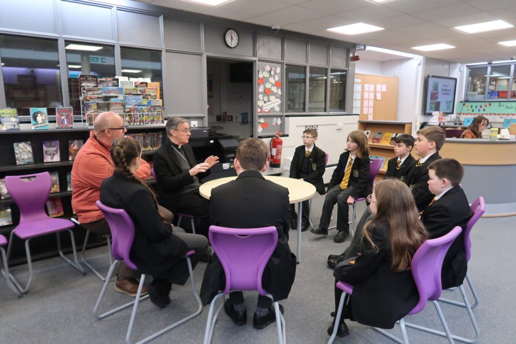 Bishop John sits in a circle with school students and a teacher in discussion in the library