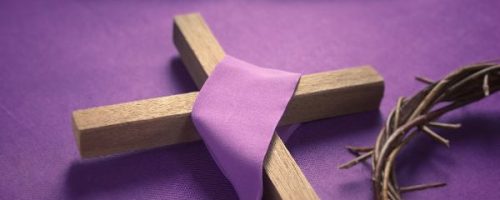 A wooden cross, draped in purple cloth, a crown of thorns, and a Bible sit on top of a purple cloth