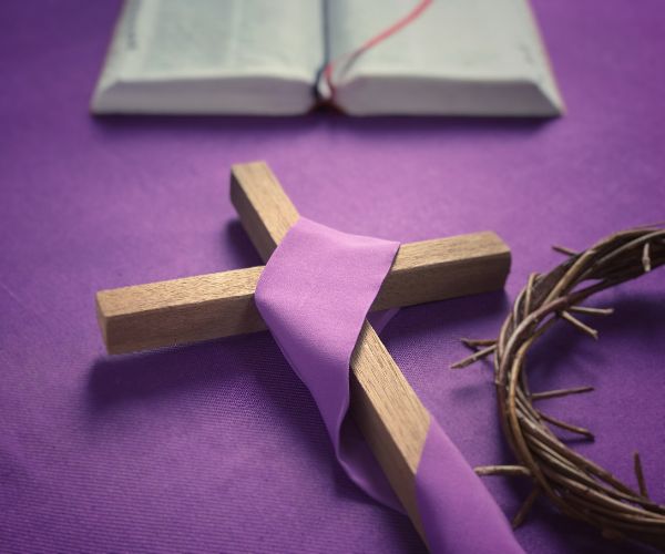 A wooden cross, draped in purple cloth, a crown of thorns, and a Bible sit on top of a purple cloth