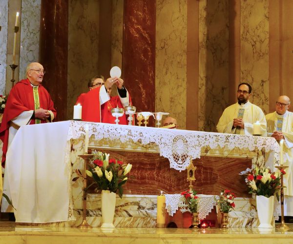 The Nuncio, dressed in red vestments, elevates the Eucharist before the altar. Bishop Brain is to one side and priests are lined up at the other side