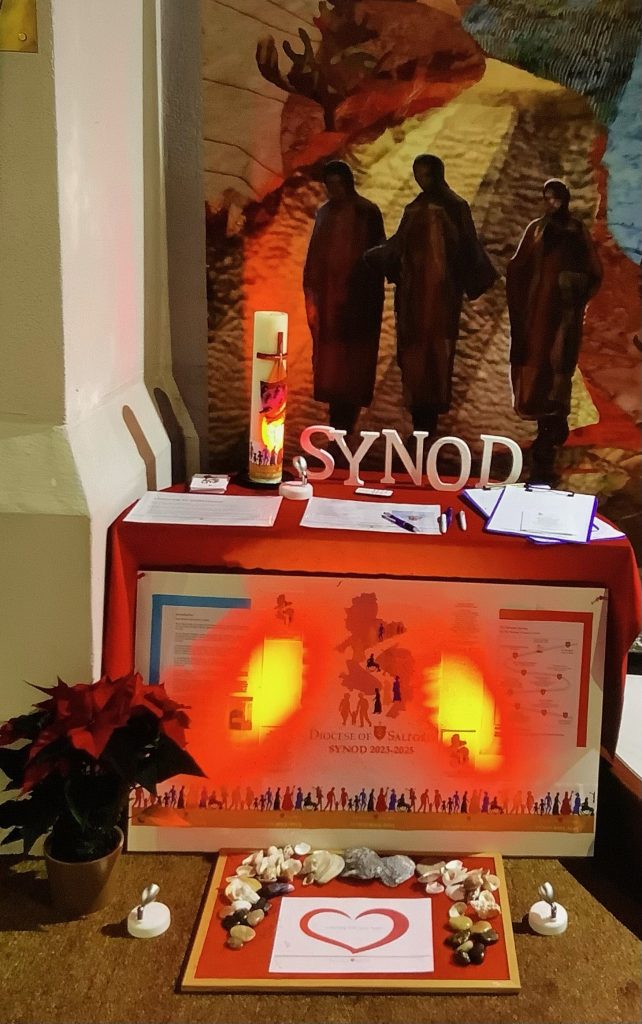 A lit pillar candle stands next to 3D letters spelling the word "Synod" on top of a table with a red table cloth, in front of which lay synod prayer cards and the synod question. A poster displaying the Synod logo was placed in front of the table, as well as further decorative features. 