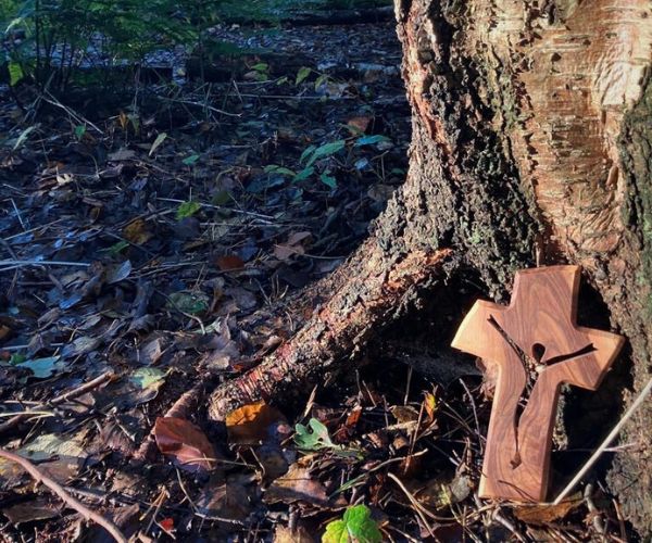A wooden cross with the figure of Christ cut out stands against the trunk of a tree in the woodland