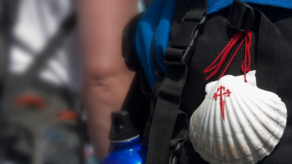 Picture shows the back of a pilgrim walking along the Camino with the iconic shell of Santiago de Compostela on their back