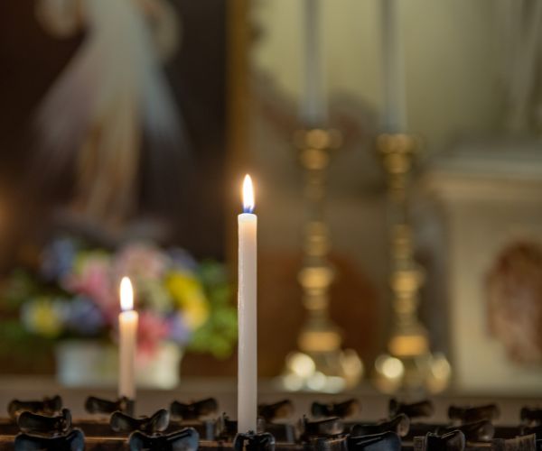 A lit white taper candle is in foreground with another a little further back and out of focused. There is the backdrop of a sanctuary and a paint of The Divine Mercy