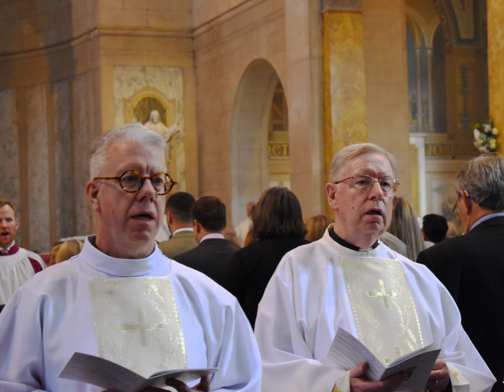 Deacon Paul and Deacon Stephen walk side by side in white vestments against the golden backdrop of St Patrick's