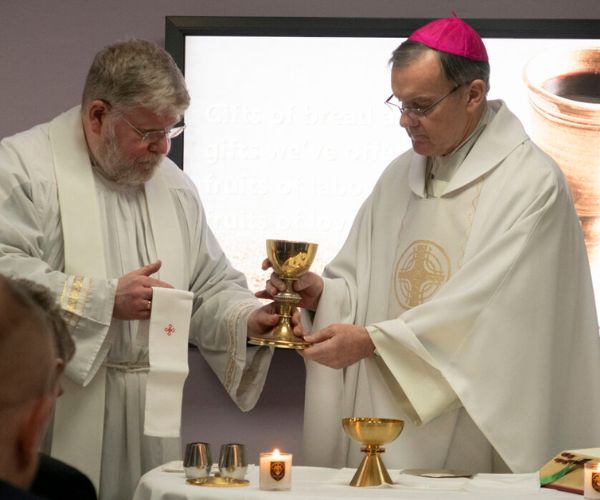 Fr Francis (left), dressed in white vestments, passes a golden chalice to Bishop John (right), also dressed in vestments, as they concelebrate Mass