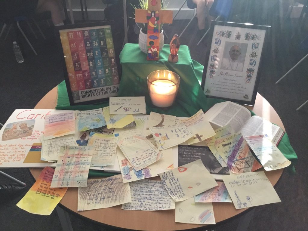 A table display featuring the colourful pledges of the pupils, a framed photo of Pope Francis, a wooden cross, and a lit candle