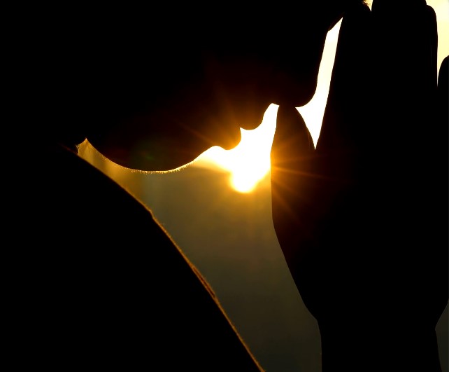 Silhouette of a person with their head bowed in prayer and their hands placed together and touching their forehead. The light of the sun shines through from the background