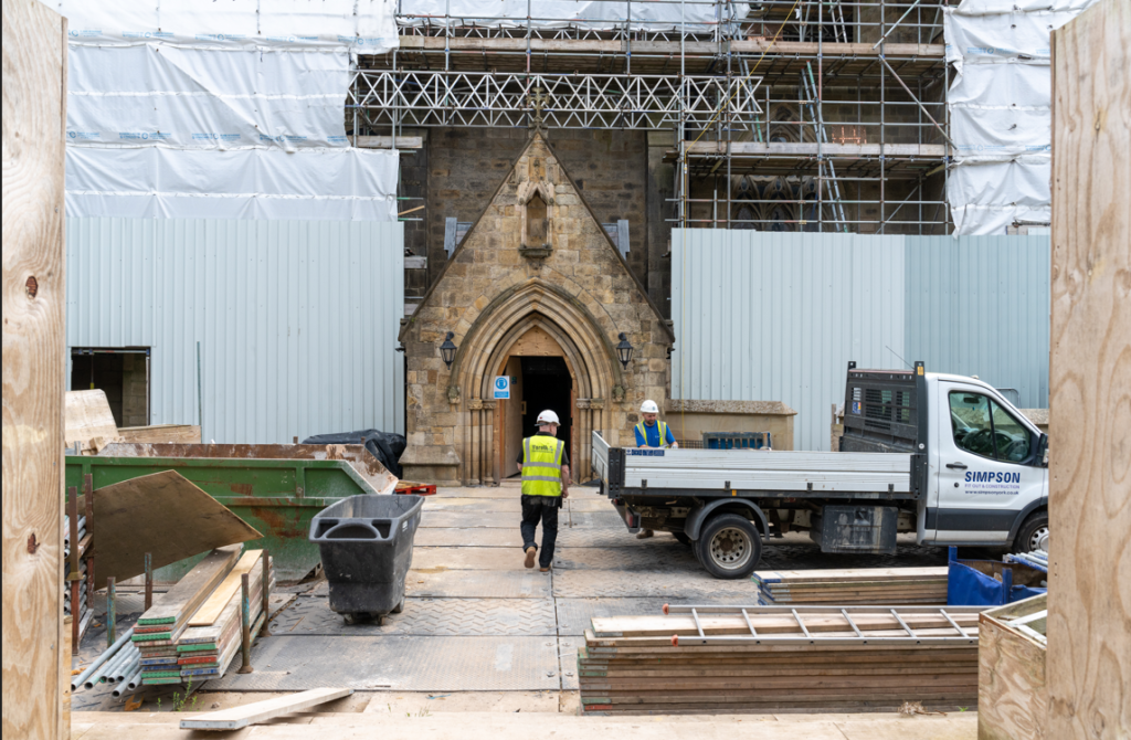 A worker in hi-vis and a hard hat walks through the piazza, past a van and building materials, into the scaffolded cathedral